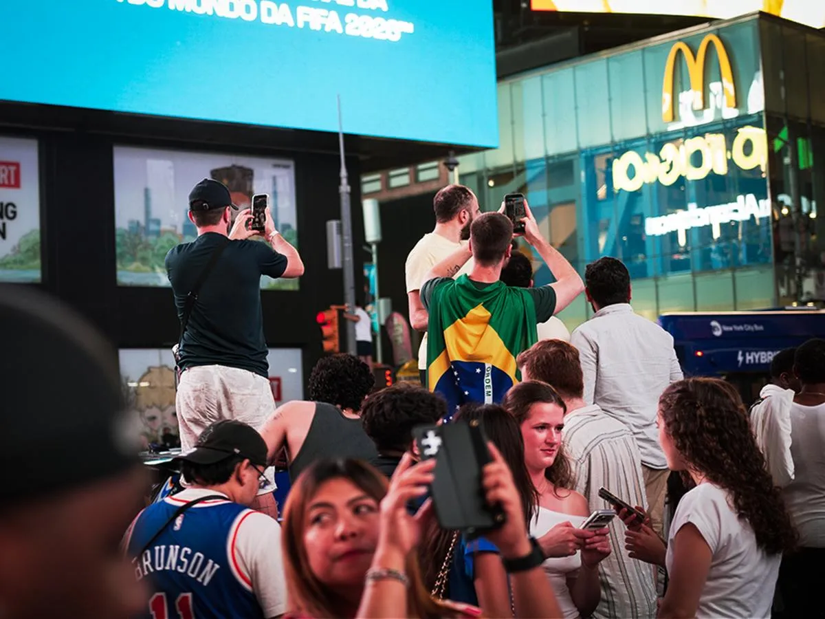 Fan draped in Brazilian flag filming Times Square with smartphone, FIFA 2026 screen visible in background with McDonald's signage