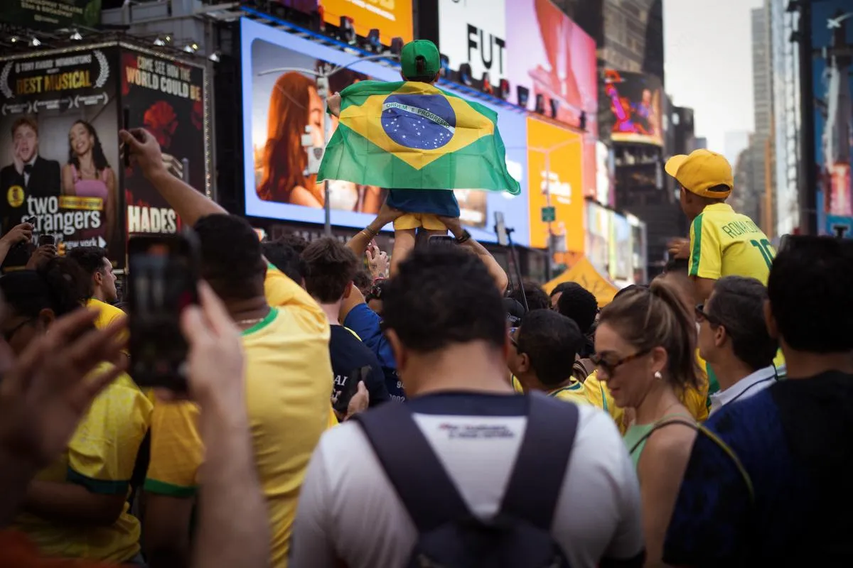 Child held high on shoulders draped in Brazilian flag, surrounded by yellow-clad fans in Times Square with Broadway billboards behind