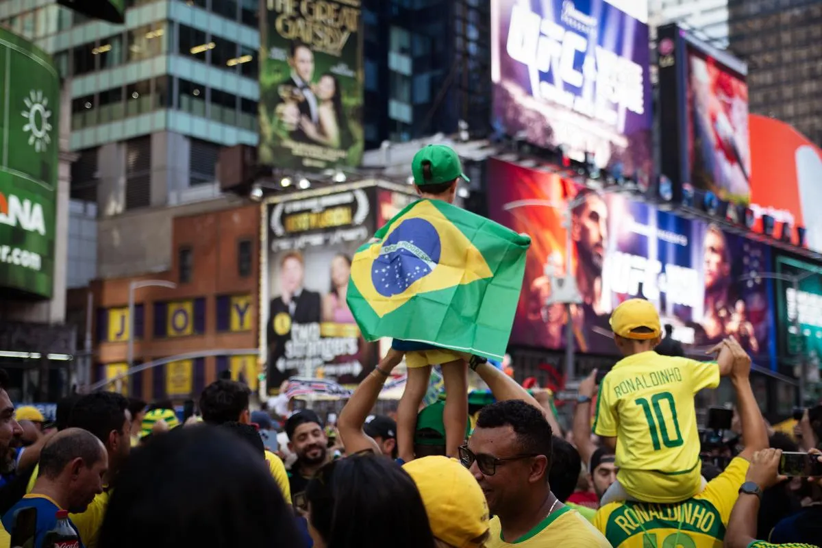 Father carrying son with Brazilian flag cape on shoulders, Ronaldinho jerseys visible in crowd, Times Square billboards including The Great Gatsby in background