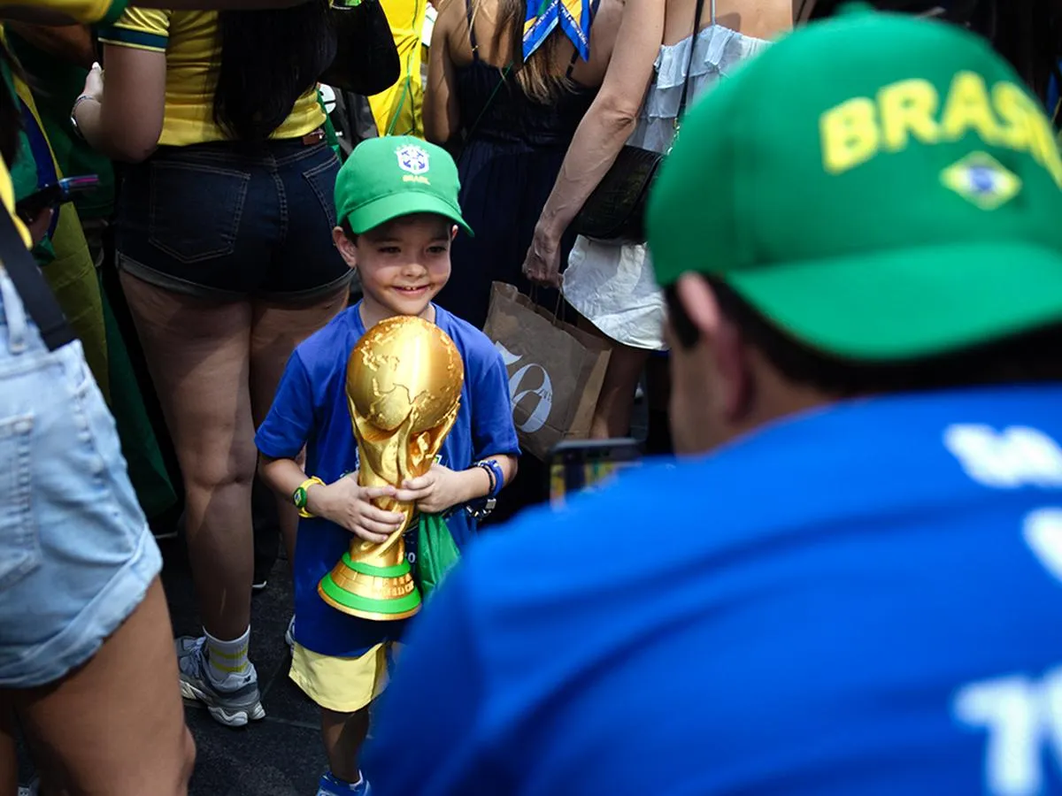 Young boy in Brazil gear holding golden World Cup trophy replica, smiling proudly while surrounded by adult fans