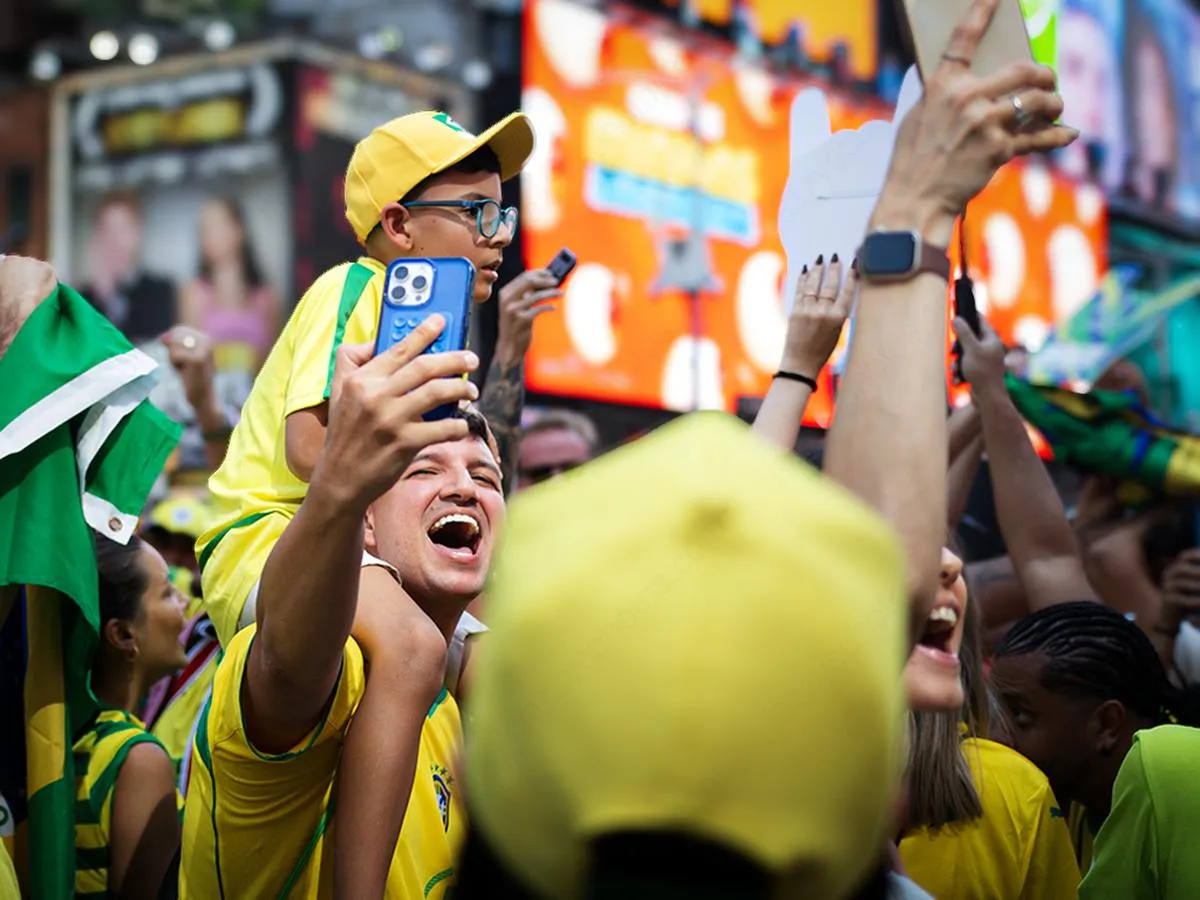 Ecstatic father screaming with joy while carrying young son on shoulders who films with smartphone, surrounded by cheering Brazilian fans