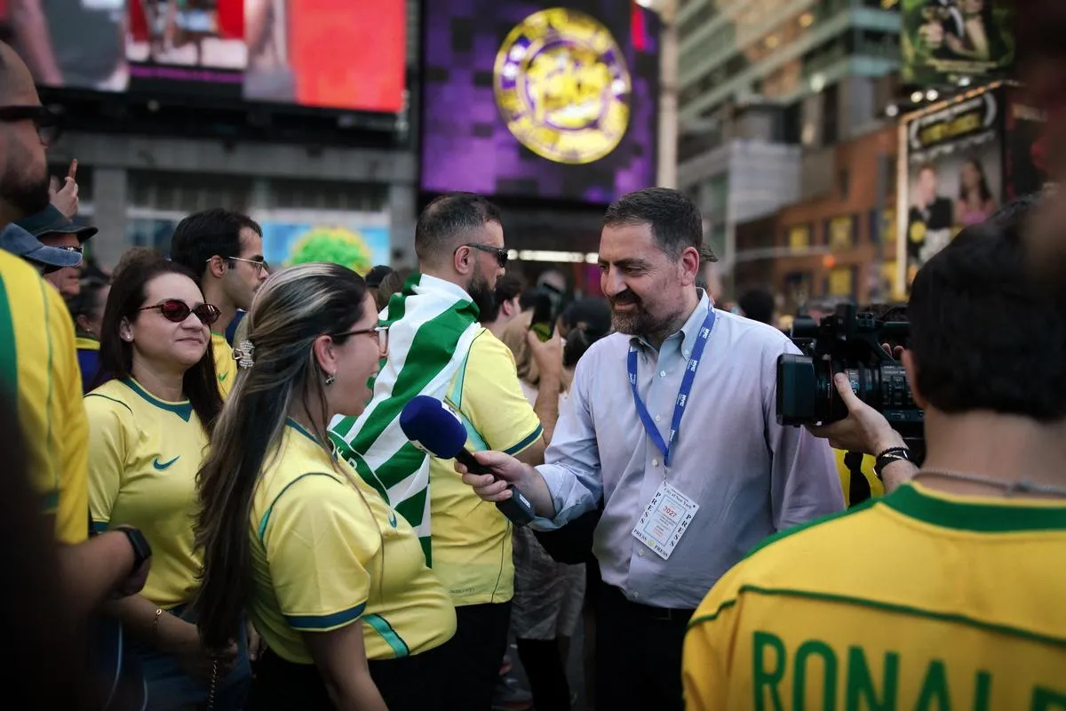 TV journalist with blue microphone interviewing smiling Brazilian fan in yellow jersey, camera crew filming in Times Square