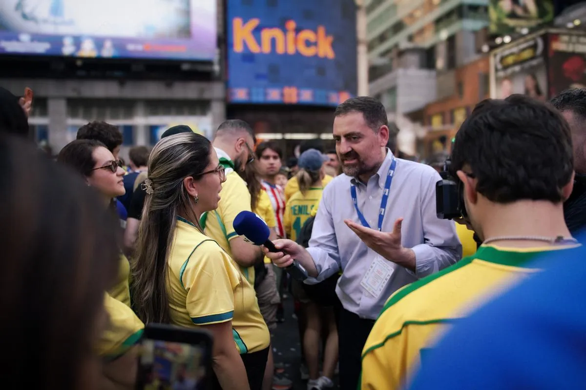 Reporter with press credentials gesturing expressively while interviewing female Brazilian fan in yellow jersey, camera crew behind