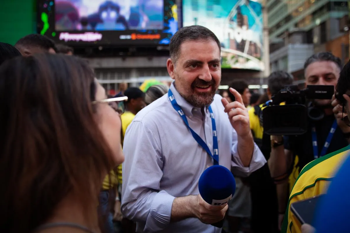 Reporter with blue microphone making emphatic point during interview, Brazilian fans in yellow jerseys surrounding him in Times Square