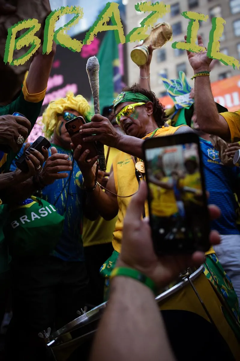 Superfan holding drum stick and World Cup trophy replica surrounded by phones filming, fan in yellow wig beside him, BRAZIL text overlay
