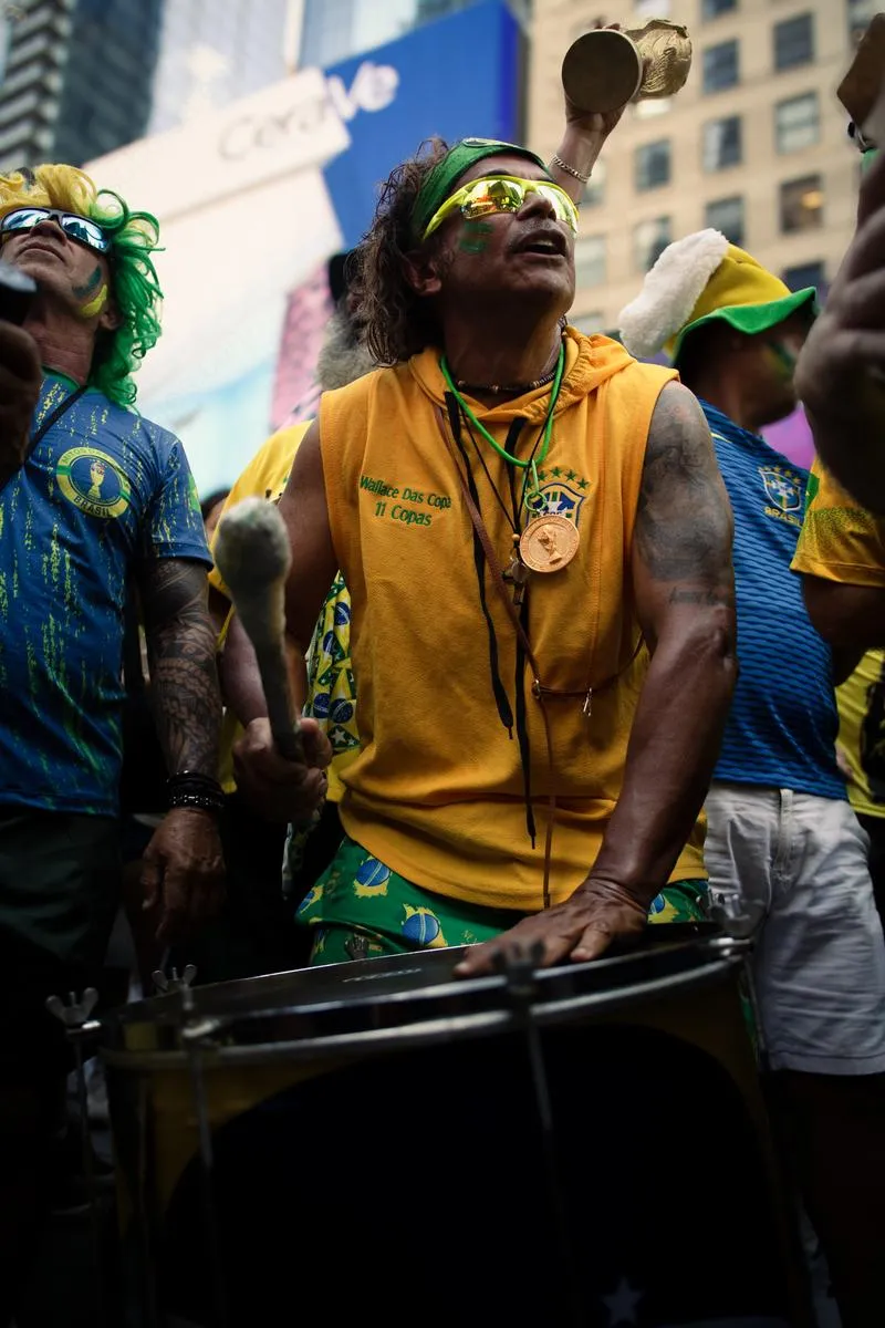Devoted Brazilian superfan playing snare drum looking skyward, wearing yellow sleeveless shirt with Brazil crest and green headband, colorful-wigged fan beside him