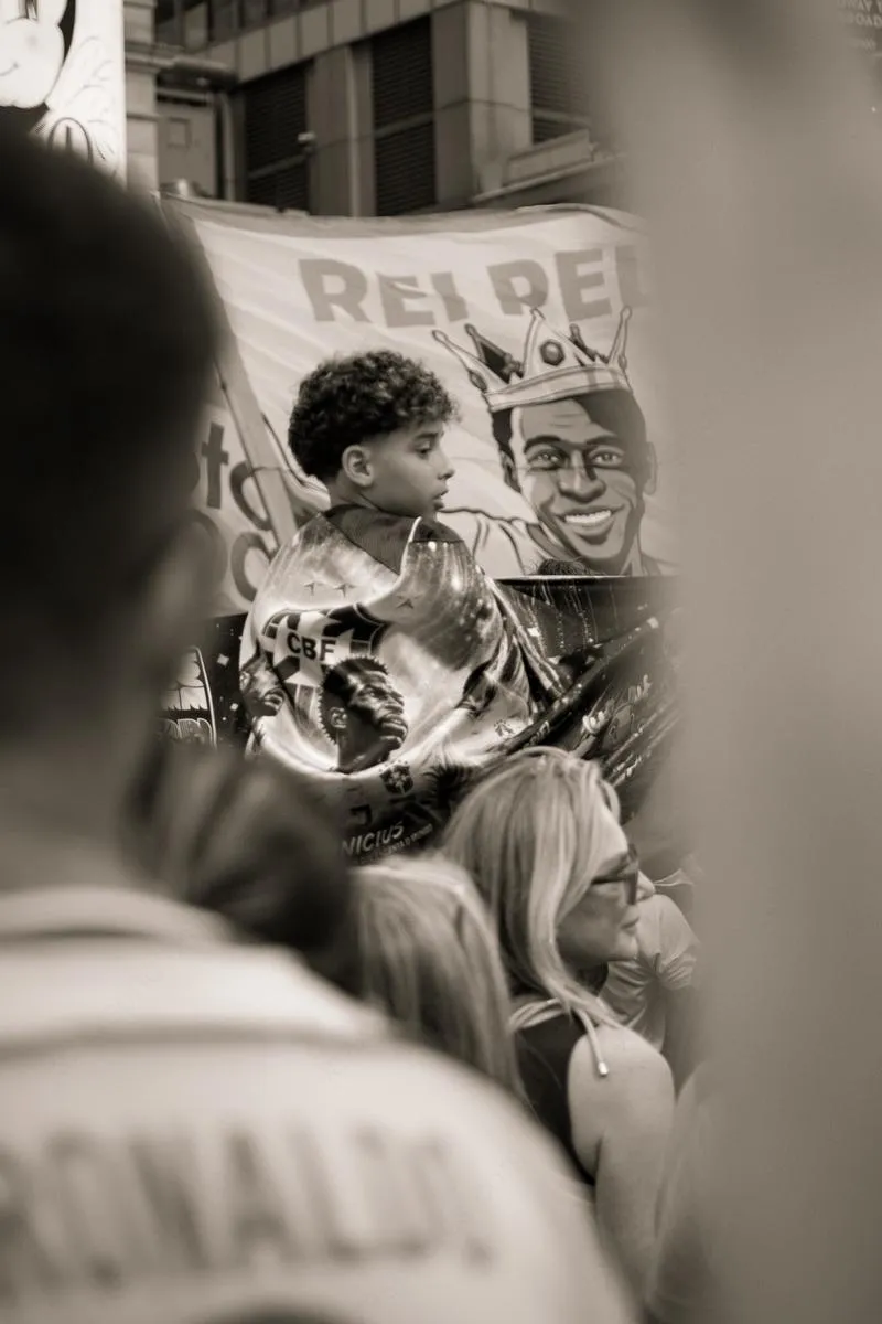 Black and white photograph of young boy on shoulders holding Brazil balloon, Rei Pele banner visible behind, Ronaldo jersey in foreground