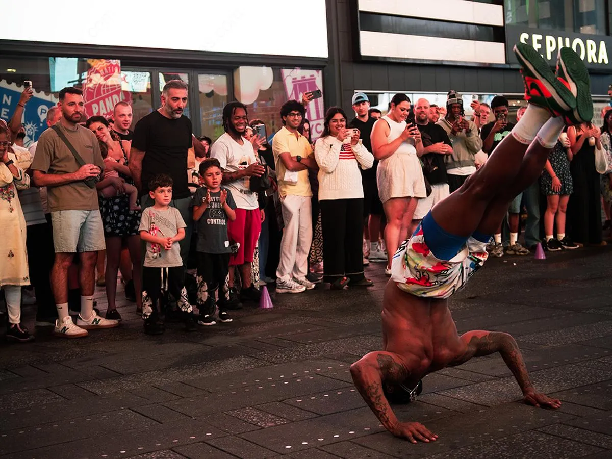 Breakdancer performing a one-handed freeze while a diverse crowd watches and records on their phones in Times Square