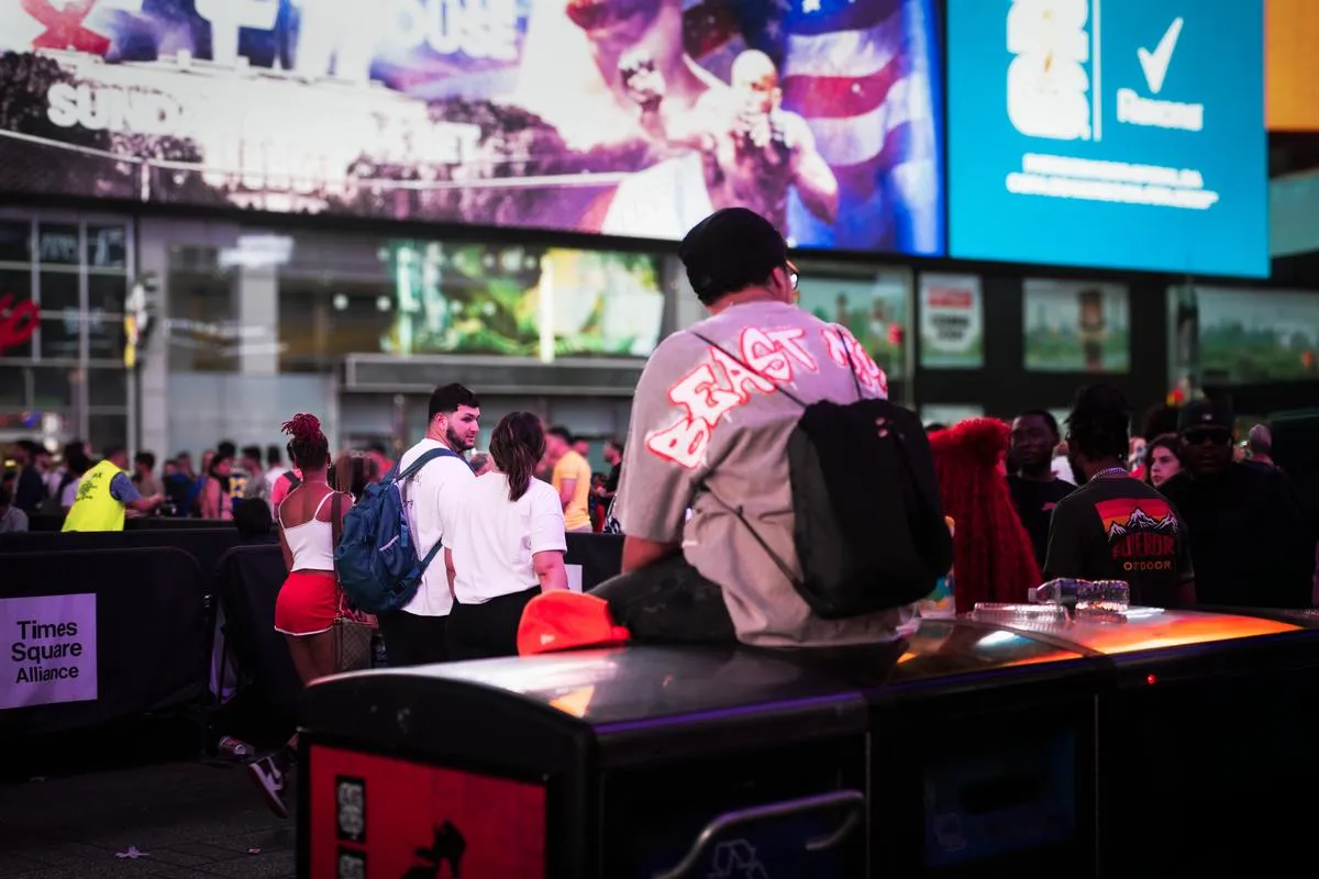 Man in East Coast shirt sits on equipment cases watching crowds gather in Times Square at night with billboards glowing