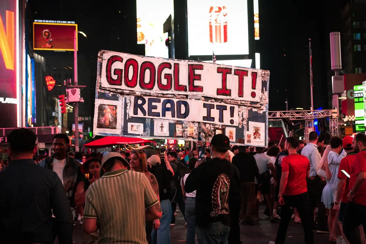 Person holding large Google It Read It sign above crowd in nighttime Times Square with billboards illuminating the scene