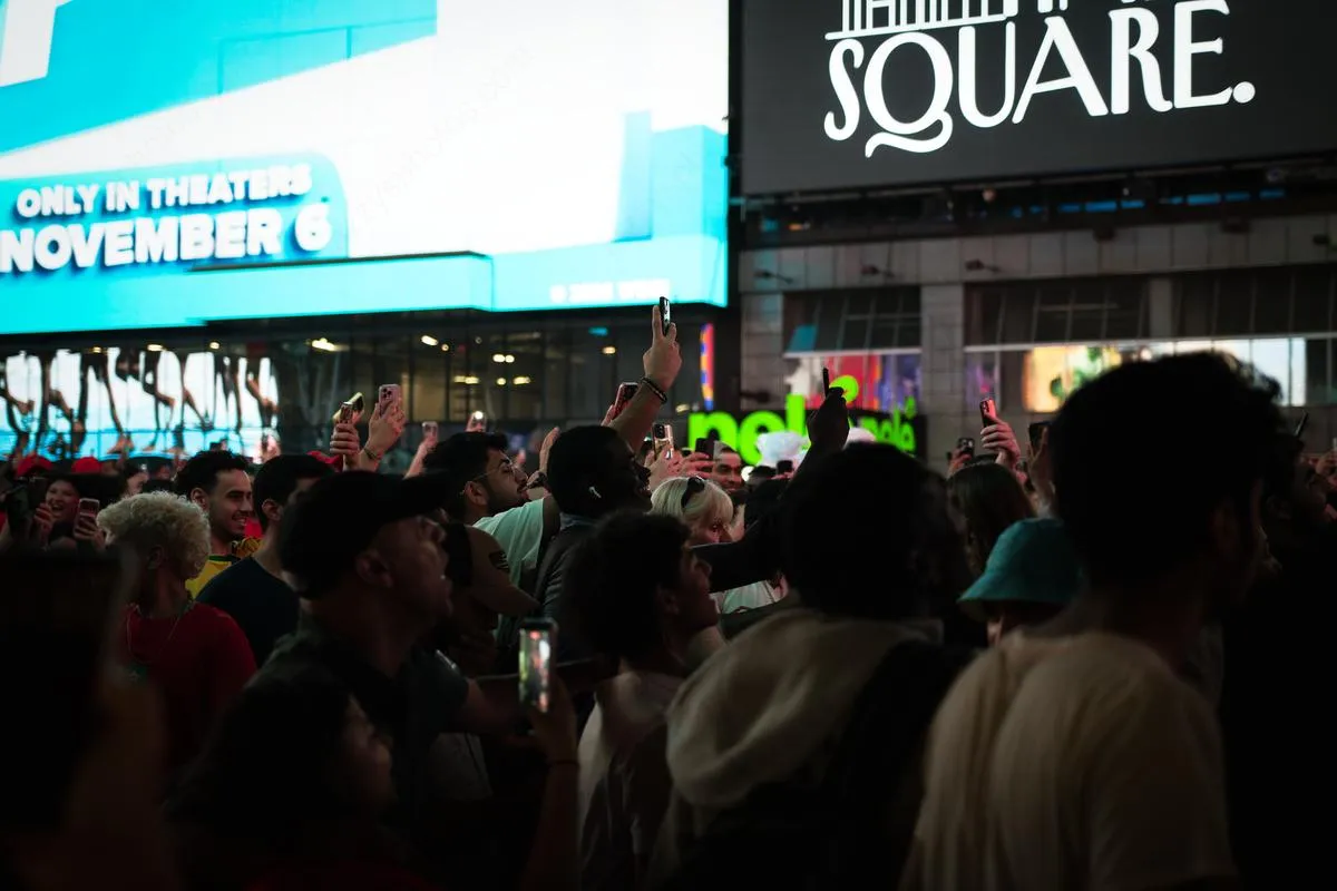 Dense crowd of fans holding up phones to record in Times Square with illuminated billboards including Square sign visible