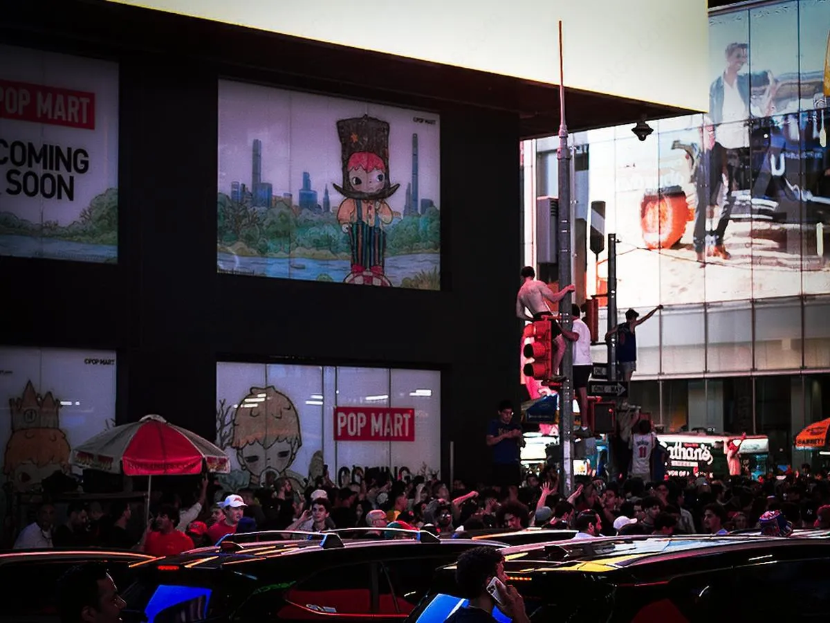 Wide view of Times Square celebration with Pop Mart store visible and fans climbing poles amid massive crowd