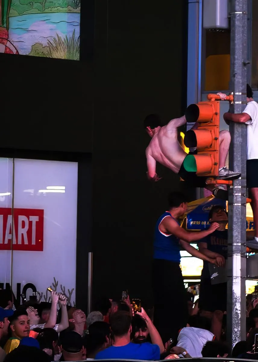 Fan hanging dramatically from traffic light with legs extended as another fan clings to pole in Times Square
