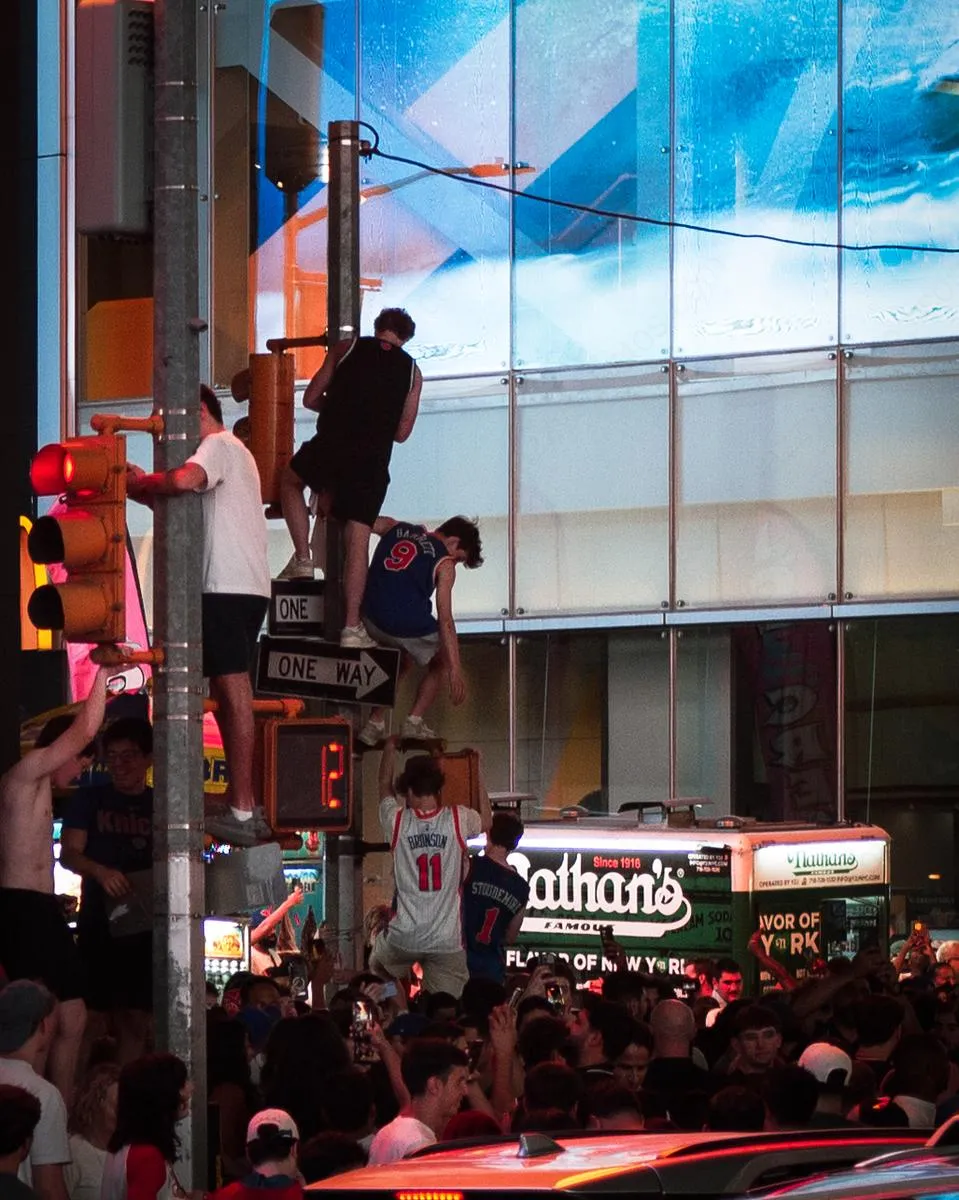 Fans climbing traffic poles with Knicks jerseys visible and Nathan's Famous hot dog truck in background