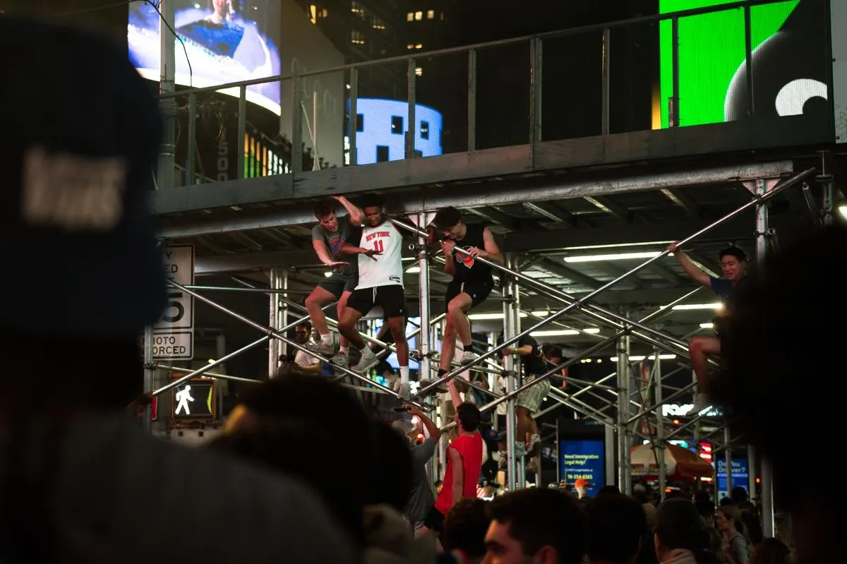 Young men in Knicks jerseys hanging from scaffolding structure in Times Square with billboards behind them