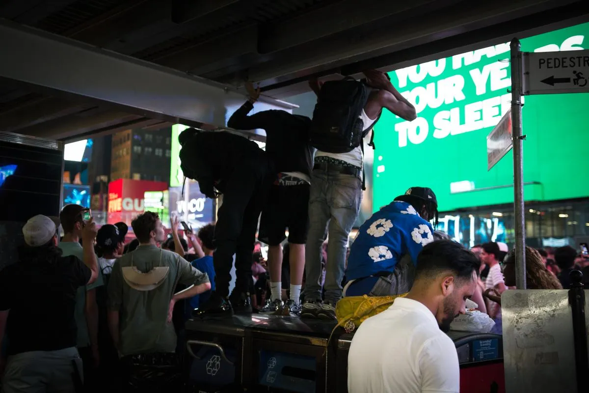 Young men standing on trash cans and hanging from awning in Times Square with Five Guys sign visible