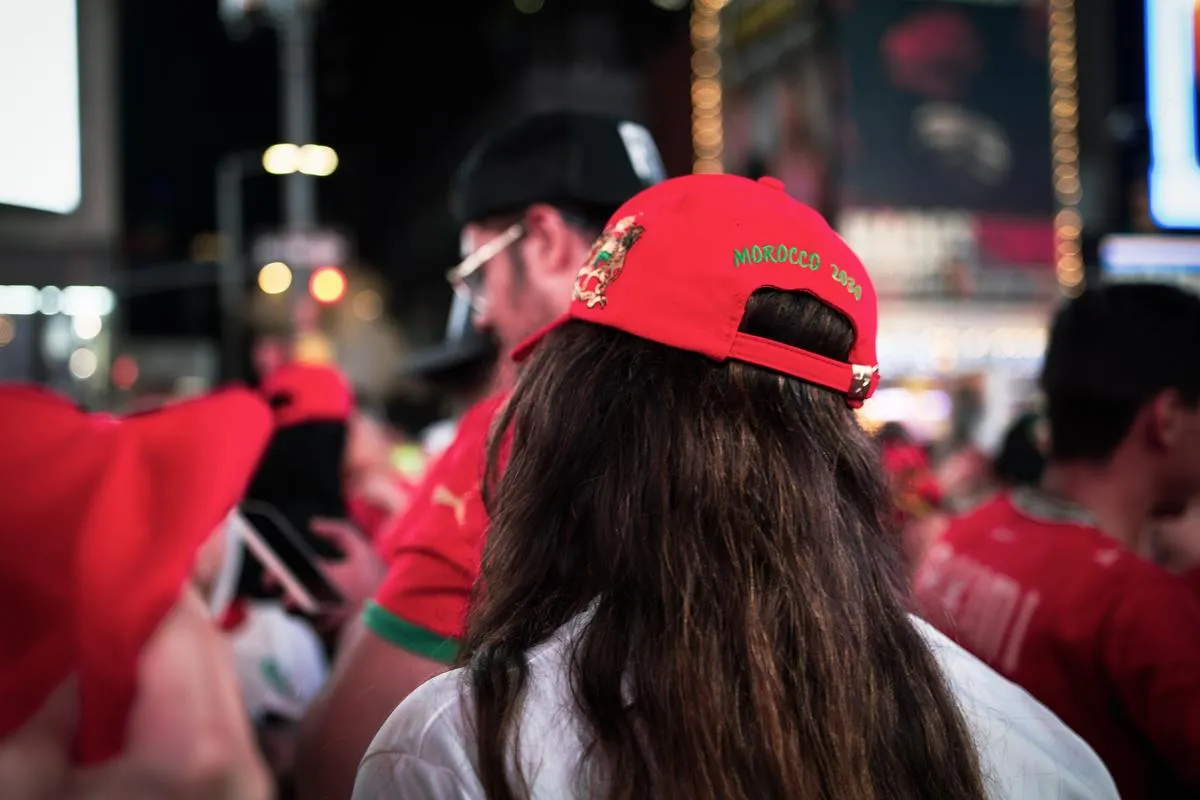 Back view of a fan wearing a red Morocco 2026 cap with long brown hair, surrounded by other supporters in red at a Times Square celebration