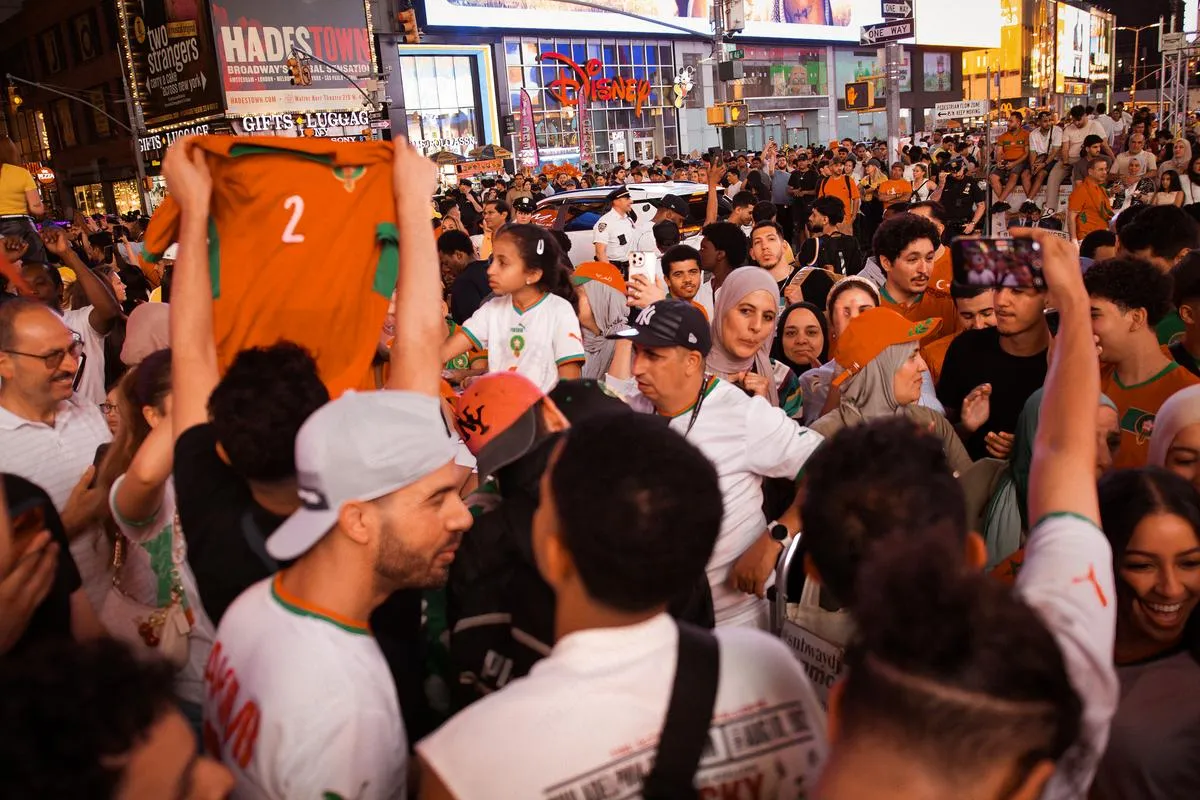 Crowded Times Square street scene with Morocco fans holding up jerseys, Broadway billboards including Hadestown visible in background