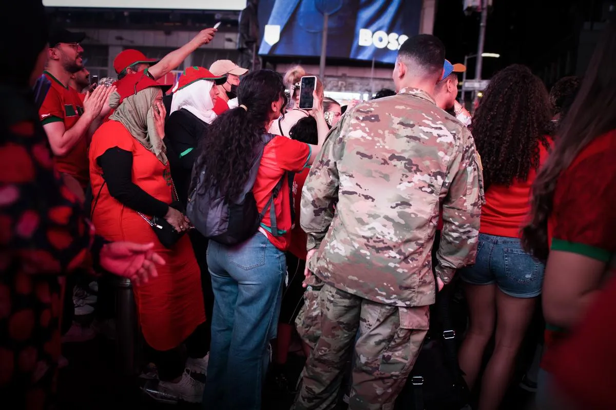 Diverse group of Morocco fans including women in hijabs and a person in military camouflage uniform, capturing the moment on phones in Times Square