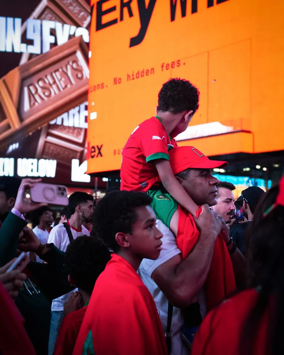Young boy in Morocco jersey sitting on his father's shoulders amid a crowd of fans, Times Square billboards glowing orange behind them