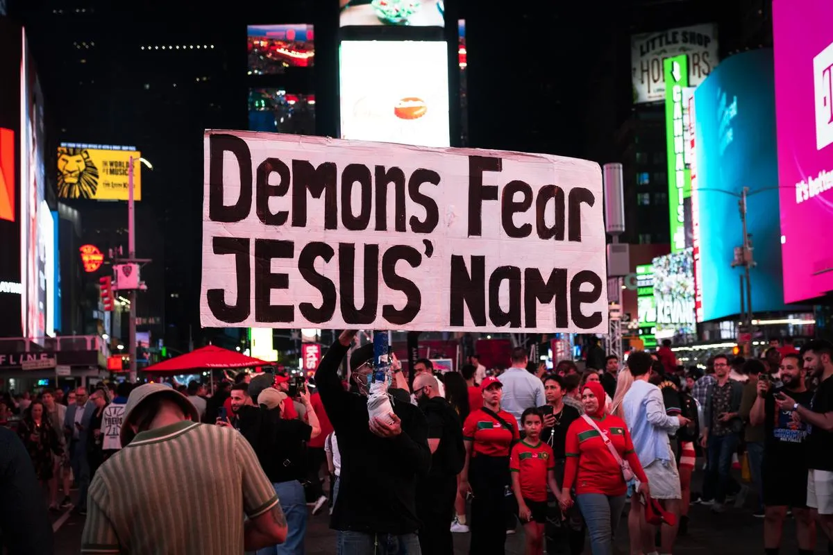Street preacher holding a large sign above the Times Square crowd with Morocco supporters in red visible below, neon lights of Broadway in background