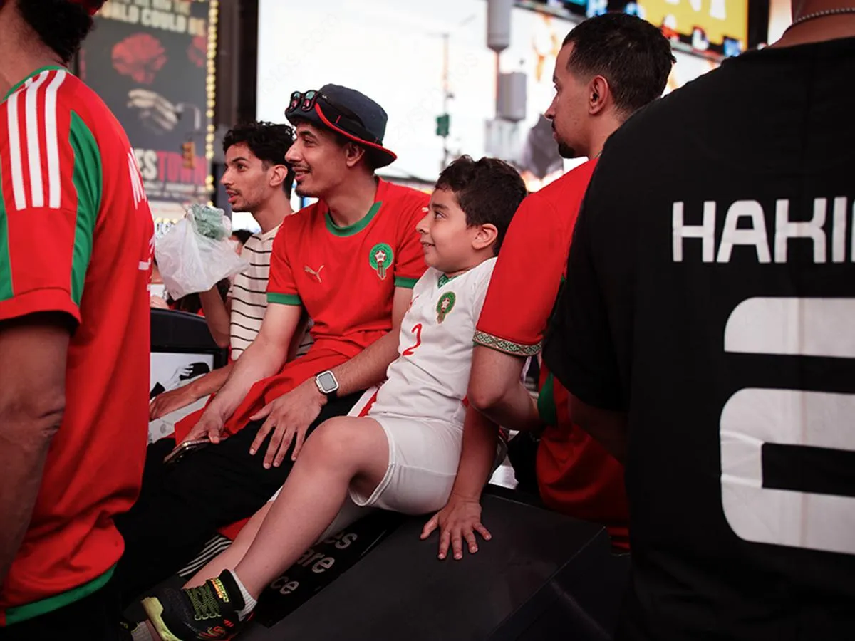 Young boy in white Morocco jersey with Hakimi name visible on nearby fan's shirt, surrounded by supporters in red and green at Times Square