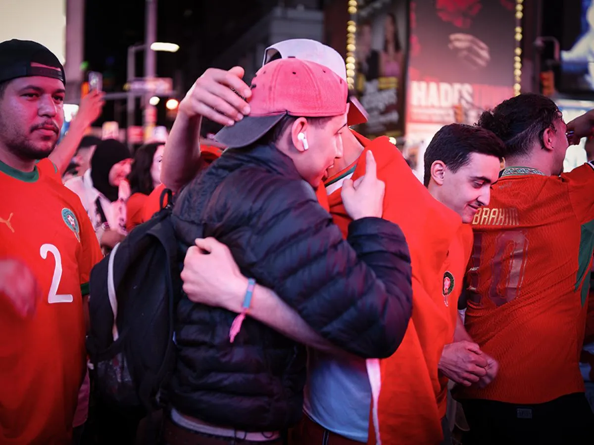 Two fans embracing emotionally amid the crowd, one in dark jacket and red cap, surrounded by Morocco supporters in red jerseys at night