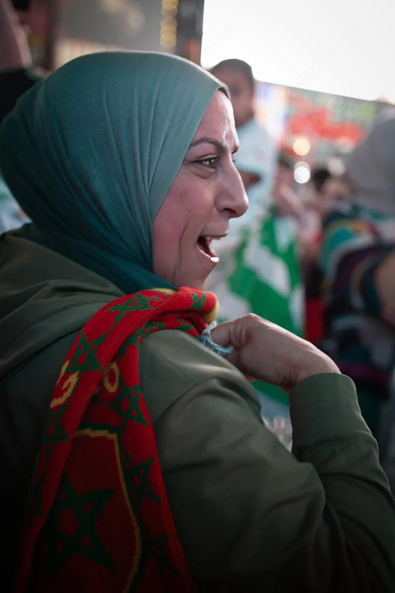 Close-up profile portrait of a woman in teal hijab with traditional red and green Morocco scarf, laughing joyfully amid celebration