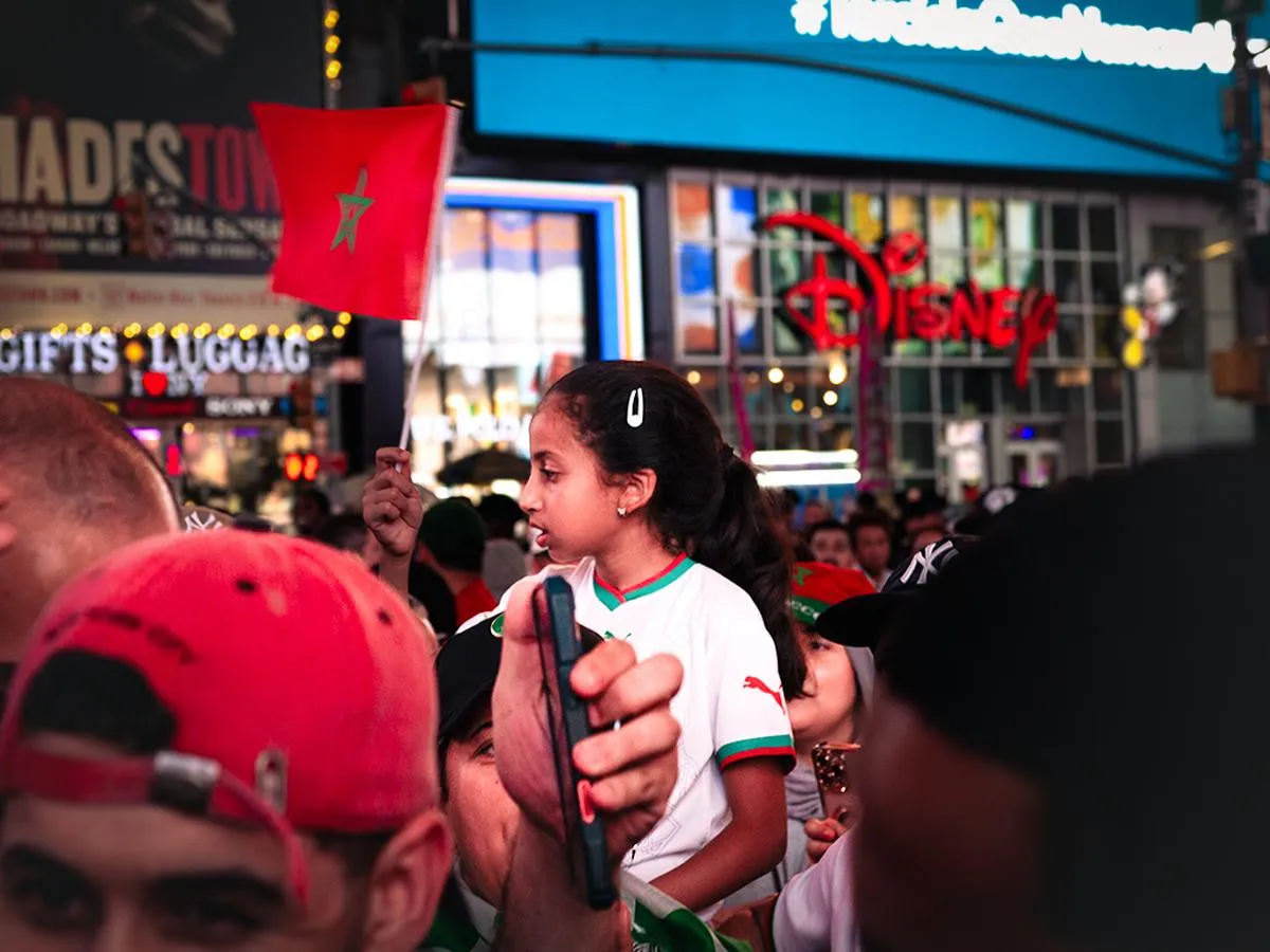 Young girl in white Morocco jersey waving a small red Moroccan flag in Times Square, Disney Store and Hadestown marquee visible in background